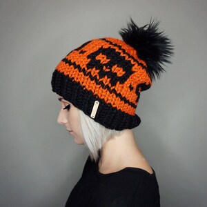 May include: A black and orange knit beanie with a black faux fur pom pom. The beanie has a geometric pattern.