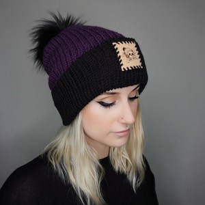 May include: A black and purple knit beanie with a faux fur pom pom and a leather patch with a skull and crossbones design.