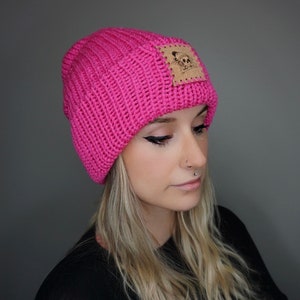May include: A pink knitted beanie with a brown leather patch featuring a skull logo.
