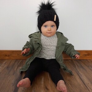 May include: A baby wearing a black knit beanie with a fluffy pom pom, a green jacket, and black pants. The beanie has a small wooden tag attached to it.