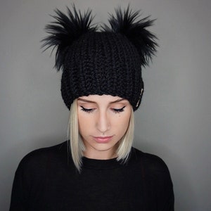 May include: A black knit beanie with two large, fluffy black pom-poms. The hat has a ribbed texture and a small gold accent. The beanie is a winter accessory.