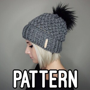 May include: A gray crocheted beanie with a black pom pom. The beanie has a textured stitch pattern and a small brown label with the word "PATTERN" on it.