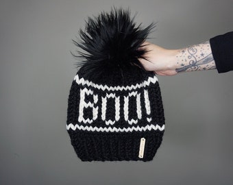 Op de afbeelding: Zwarte gebreide beanie met een witte "Boo!"-print en een zwarte nep bont pompon.