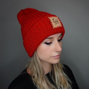 May include: A red knitted beanie with a brown leather patch featuring a skull graphic. The beanie is folded down and has a ribbed texture.