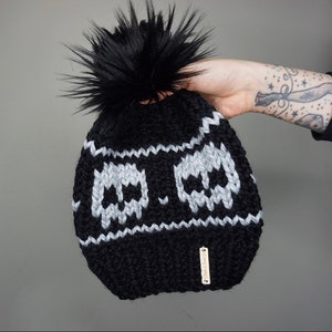 May include: Black knit beanie with a white skull pattern and a black faux fur pom pom. The beanie has a small brown tag with the text "Handmade" on it.