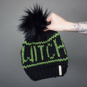 May include: Black knit beanie with a green "WITCH" design and a black faux fur pom pom.