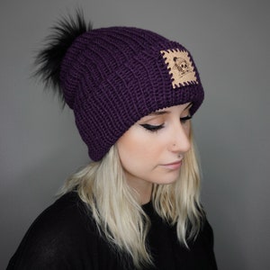 May include: Purple knitted beanie with a black faux fur pom pom and a brown leather patch with a skull design.