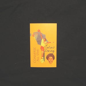 May include: An orange Converse All Star promotional card featuring a basketball player in action. The card includes the text "Julius Erving" and a portrait of the player. The background is a black surface.