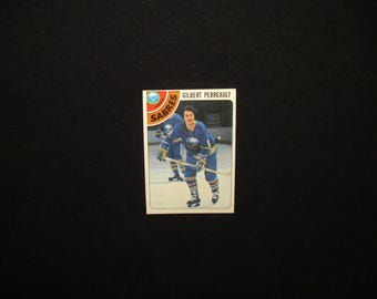 May include: A vintage hockey trading card featuring Gilbert Perreault of the Buffalo Sabres. The card shows Perreault in his blue and gold uniform, holding a hockey stick. The Sabres logo is visible in the upper left corner. The card is rectangular with a white border.