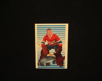 May include: A vintage hockey card featuring a goalie in a red, white, and blue uniform with a blue and white striped background. The card has a black and white photo of the goalie in action, with a black and white puck in front of him. The card has the player's name, "Frank L. Clair", printed in black.