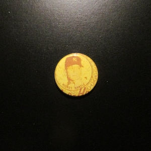 May include: A vintage yellow button featuring a portrait of Mickey Mantle wearing a baseball cap. The button has a red and yellow color scheme, with the text "Mickey Mantle" and "NY" visible.