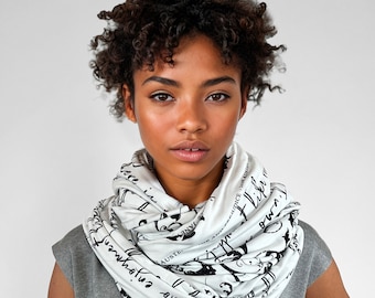 May include: A white infinity scarf featuring black printed text and illustrations. The fabric is draped in multiple loops around the neck, displaying cursive lettering and graphic elements. The scarf is worn over a plain gray short-sleeved top.