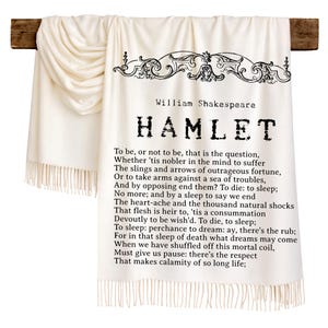 Könnte beinhalten: Eine cremeweiße Wohndecke mit einem schwarzen Aufdruck eines Auszugs aus William Shakespeares Hamlet, einschließlich des berühmten Monologs beginnend mit To be, or not to be. Die Decke hat eine dekorative Schmuckbordüre und Fransen am unteren Rand.
