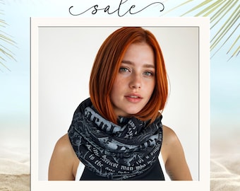 May include: A black infinity scarf featuring a white printed pattern of gothic-style text and illustrations. The scarf is worn around the neck, displaying a soft, draped fabric texture with visible white lettering against the dark background.