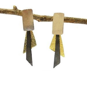 Könnte beinhalten: Ein Paar abstrakte Ohrringe mit rechteckigem Oberteil und geschichtetem Design. Das Oberteil ist hellbraun, mit goldenen und schwarzen Akzenten. Die Ohrringe hängen an einem braunen Ast vor weißem Hintergrund.