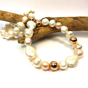 May include: A pearl necklace with a mix of white, blush pink, and rose gold beads. The necklace features a variety of pearl shapes, including round and baroque, and is accented with metallic rose gold spheres. The necklace is displayed on a piece of driftwood.