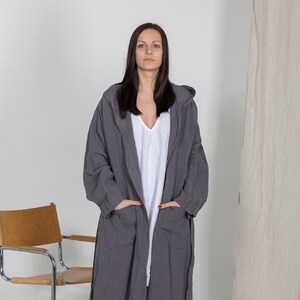 May include: A long, gray hooded robe with a tie belt. The robe is made of a soft, textured fabric. The model is wearing the robe with a white shirt underneath.