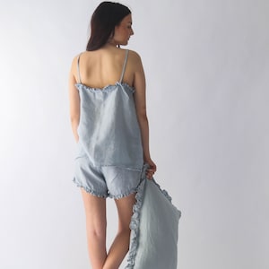Puede incluir: Un conjunto de pijama de lino azul claro con ribete de volantes. El conjunto incluye una camiseta sin mangas y pantalones cortos. La modelo lleva el conjunto y sostiene una almohada a juego.