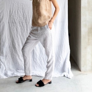 May include: A woman wearing a light brown sleeveless top and white linen pants. She is wearing black heeled sandals with a bow detail.