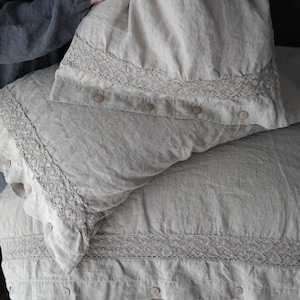 May include: Beige linen bedding set featuring a duvet cover and pillowcases. The duvet cover and pillowcases have a decorative lace trim along the edges and are secured with neutral-colored buttons. The fabric has a slightly wrinkled texture.