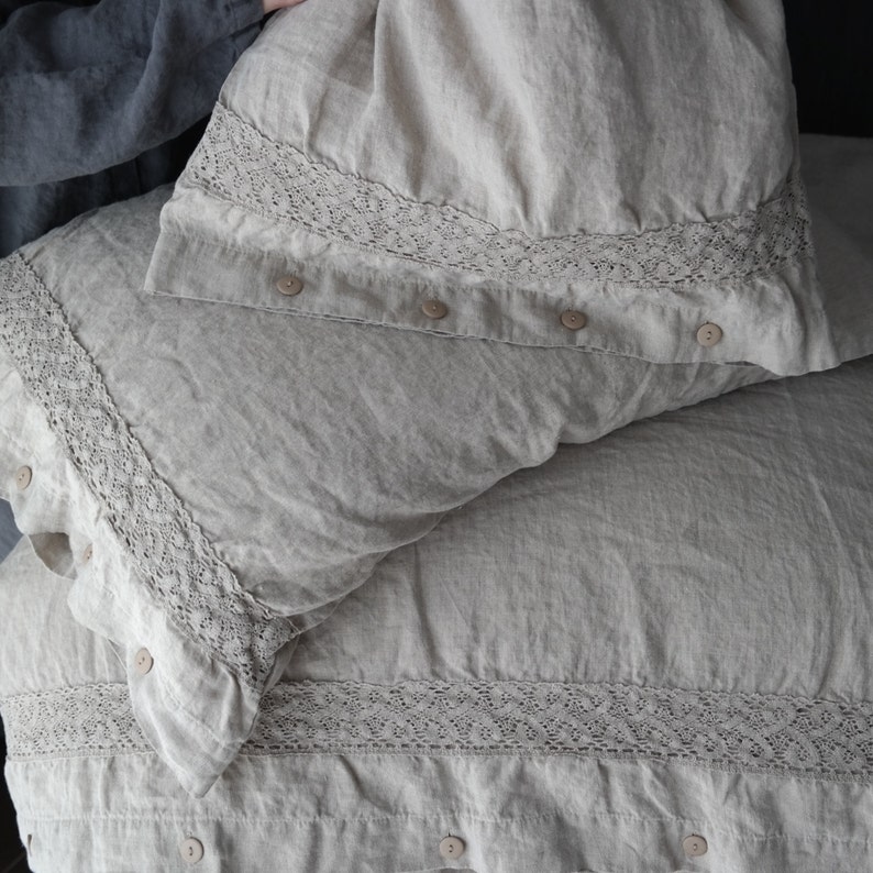 May include: Beige linen bedding set featuring a duvet cover and pillowcases. The duvet cover and pillowcases have a decorative lace trim along the edges and are secured with neutral-colored buttons. The fabric has a slightly wrinkled texture.
