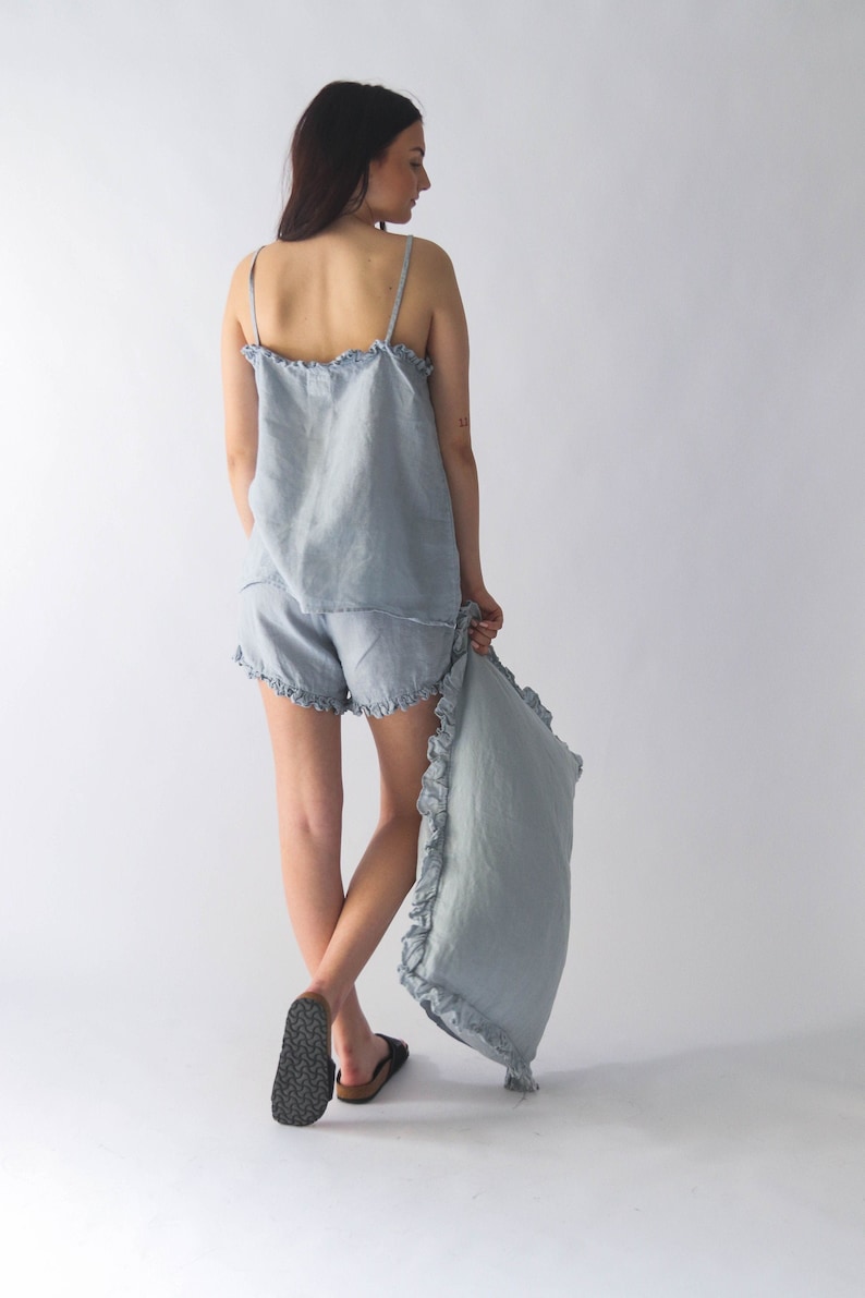 Puede incluir: Un conjunto de pijama de lino azul claro con ribete de volantes. El conjunto incluye una camiseta sin mangas y pantalones cortos. La modelo sostiene una funda de almohada a juego.