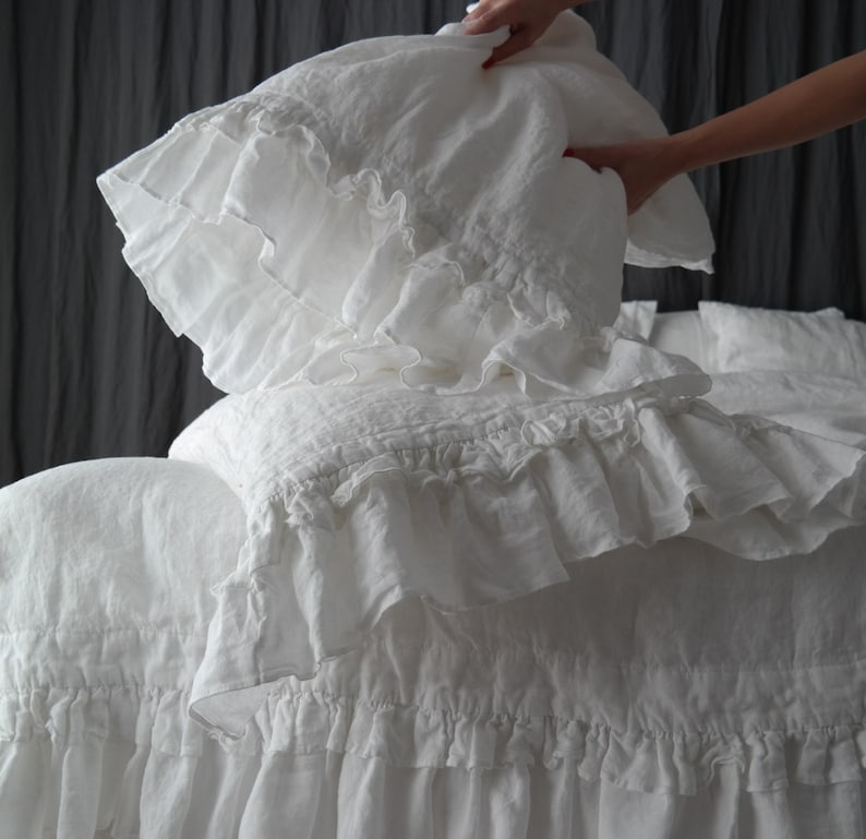 May include: White linen bedding with ruffled edges. The bedding is arranged on a bed with a white pillow and a white throw pillow.