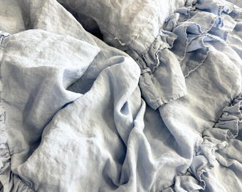 May include: A light blue linen bedding set, featuring a ruffled duvet cover and pillow. The fabric appears soft and slightly wrinkled, with a natural, textured look. The bedding is arranged on a bed, creating a cozy and inviting atmosphere.