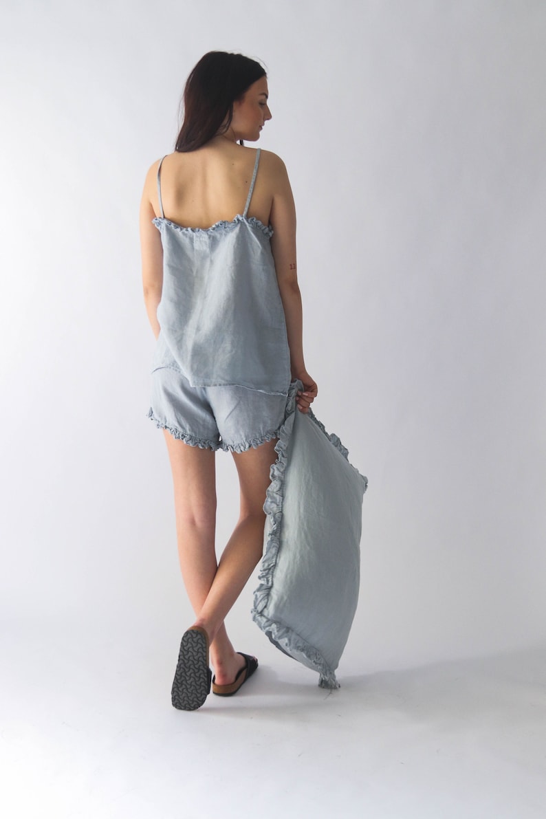 Puede incluir: Un conjunto de pijama de lino azul claro con ribete de volantes. El conjunto incluye una camiseta sin mangas y pantalones cortos. La modelo lleva el conjunto y sostiene una almohada a juego.