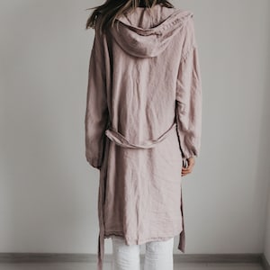K&ouml;nnte beinhalten: Eine altrosa Kapuzenjacke mit langen &Auml;rmeln und einem Bindeg&uuml;rtel, kombiniert mit einer wei&szlig;en Hose. Die Jacke hat eine lockere Passform und eine Kapuze, w&auml;hrend die wei&szlig;e Hose gerade geschnitten ist. Das Ensemble ist von hinten zu sehen.