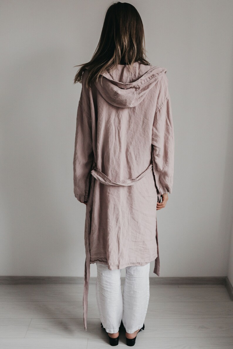 K&ouml;nnte beinhalten: Eine altrosa Kapuzenjacke mit langen &Auml;rmeln und einem Bindeg&uuml;rtel, kombiniert mit einer wei&szlig;en Hose. Die Jacke hat eine lockere Passform und eine Kapuze, w&auml;hrend die wei&szlig;e Hose gerade geschnitten ist. Das Ensemble ist von hinten zu sehen.