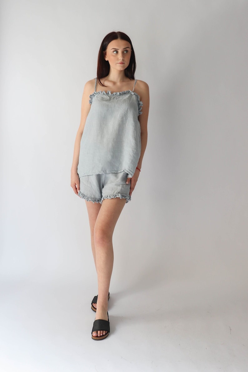 Puede incluir: Un conjunto de pijama de lino azul claro con ribete de volantes. La parte superior es un top sin mangas y los pantalones cortos son de corte holgado.