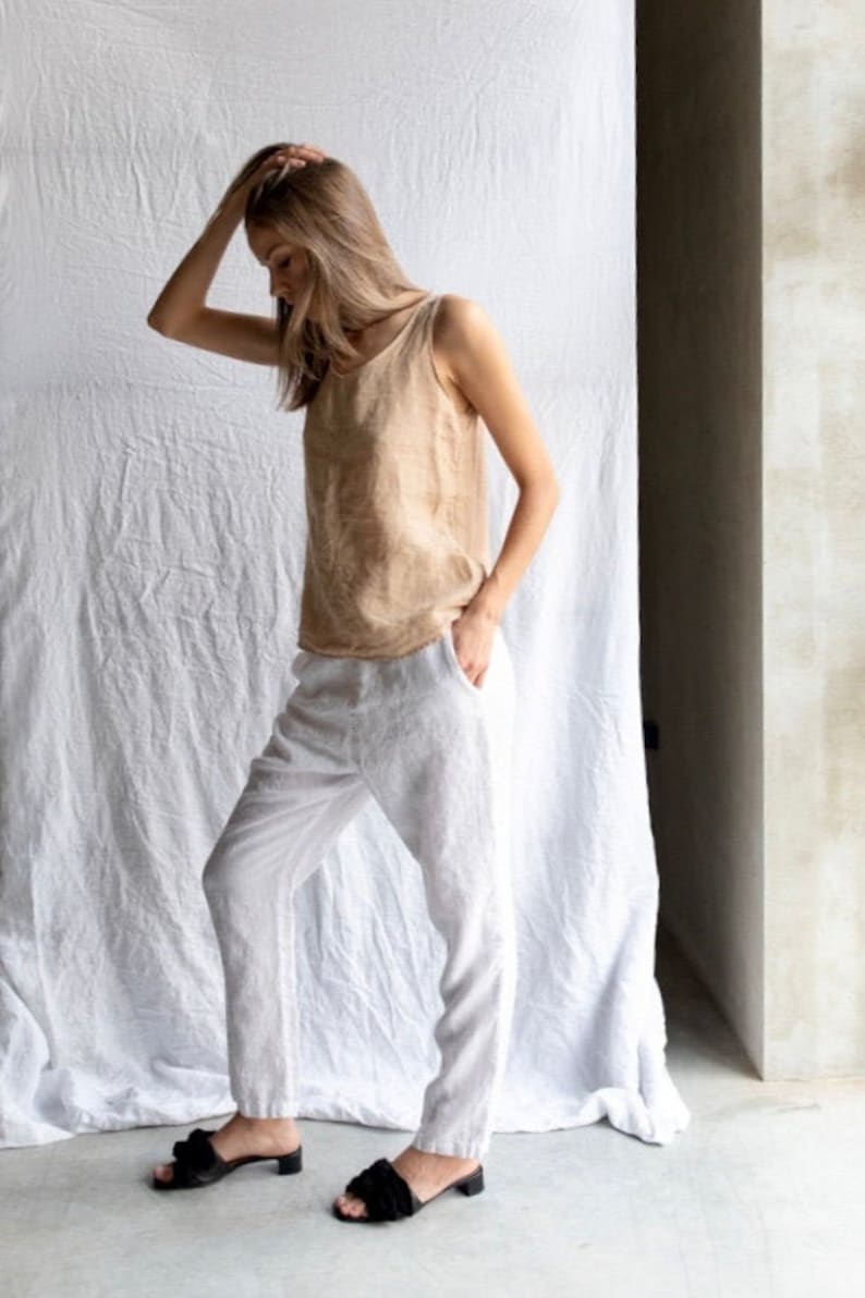 May include: A woman wearing a light brown sleeveless top and white linen pants. She is wearing black heeled sandals with a bow detail.