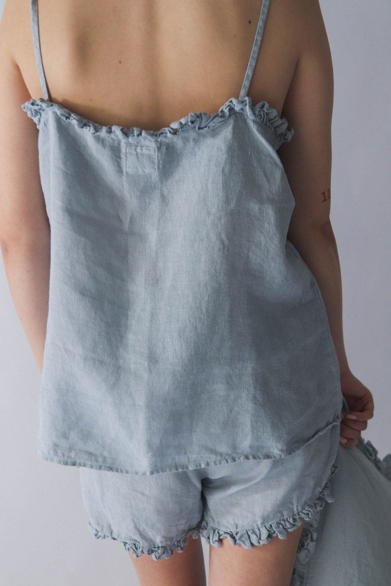 Puede incluir: Un conjunto de pijama de lino azul claro con ribete de volantes. La parte superior es un top sin mangas con tirantes finos. Los pantalones cortos tienen una pierna ancha y un dobladillo con volantes.
