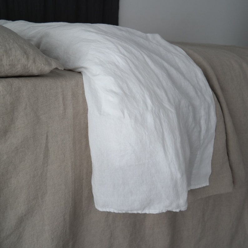 Puede incluir: Un juego de cama compuesto por una s&aacute;bana y una funda de almohada de lino beige, combinadas con una funda n&oacute;rdica de lino blanco brillante. El tejido muestra una textura natural ligeramente arrugada, ca&iacute;da sobre el borde de la cama.