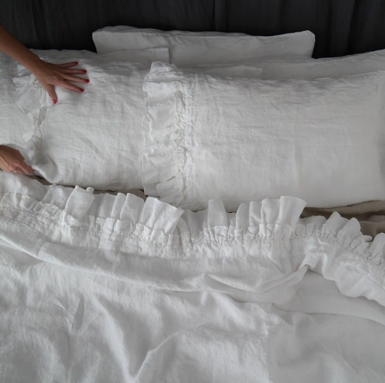 May include: White linen bedding with a ruffled edge. The bedding is made of soft, natural linen and has a relaxed, comfortable look.