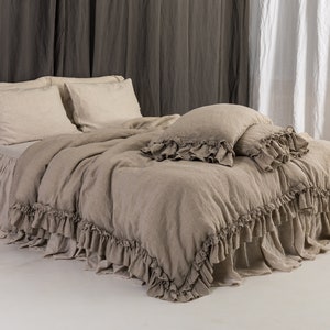 May include: A beige linen duvet cover with a ruffled edge. The duvet cover is on a bed with two beige linen pillowcases and a beige linen throw pillow. The bed is against a gray wall and a white curtain.
