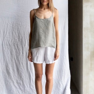 May include: A sleeveless top in a muted olive green with a subtle vertical pinstripe pattern, featuring thin spaghetti straps and a scoop neckline, paired with plain white shorts and black slide sandals with a decorative knotted detail.