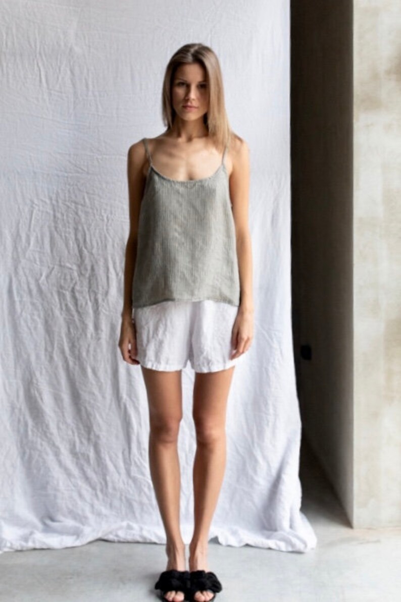 May include: A sleeveless top in a muted olive green with a subtle vertical pinstripe pattern, featuring thin spaghetti straps and a scoop neckline, paired with plain white shorts and black slide sandals with a decorative knotted detail.