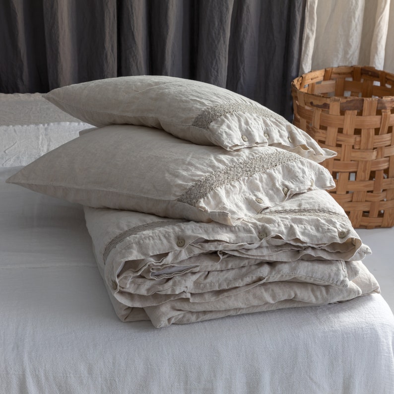May include: A stack of beige linen bedding, including two pillowcases and a folded duvet cover, with decorative trim and button closures. The bedding is arranged on a white sheet, with a woven basket in the background. The overall aesthetic is simple and natural.