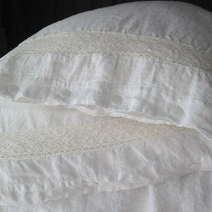 May include: Close-up of white linen pillowcases with delicate lace trim. The pillowcases have a soft, textured appearance and are secured with white buttons. The overall aesthetic is clean and elegant, suitable for bedroom decor.
