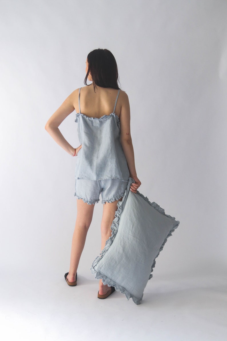 Puede incluir: Un conjunto de pijama de lino azul claro con ribete de volantes. El conjunto incluye una camiseta sin mangas y pantalones cortos. La modelo sostiene una almohada a juego con ribete de volantes.