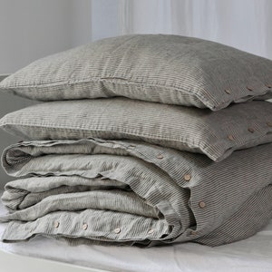 May include: A stack of gray and white striped bedding, including two pillowcases and a folded duvet cover. The duvet cover has wooden buttons along the edge. The bedding is arranged on a white surface, with a white curtain in the background.