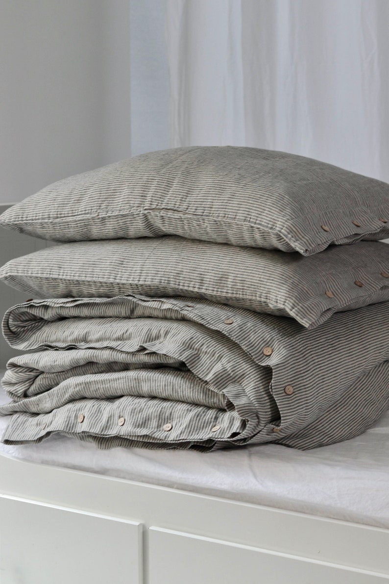 May include: A stack of gray and white striped bedding, including two pillowcases and a folded duvet cover. The duvet cover has wooden buttons along the edge. The bedding is arranged on a white surface, with a white curtain in the background.