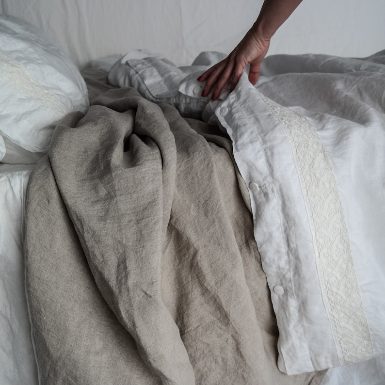 Puede incluir: Un juego de cama que incluye una funda n&oacute;rdica de lino blanco con un borde de encaje decorativo y una hilera de botones blancos, junto a una manta de lino beige liso. La tela presenta una textura de tejido natural claramente visible.
