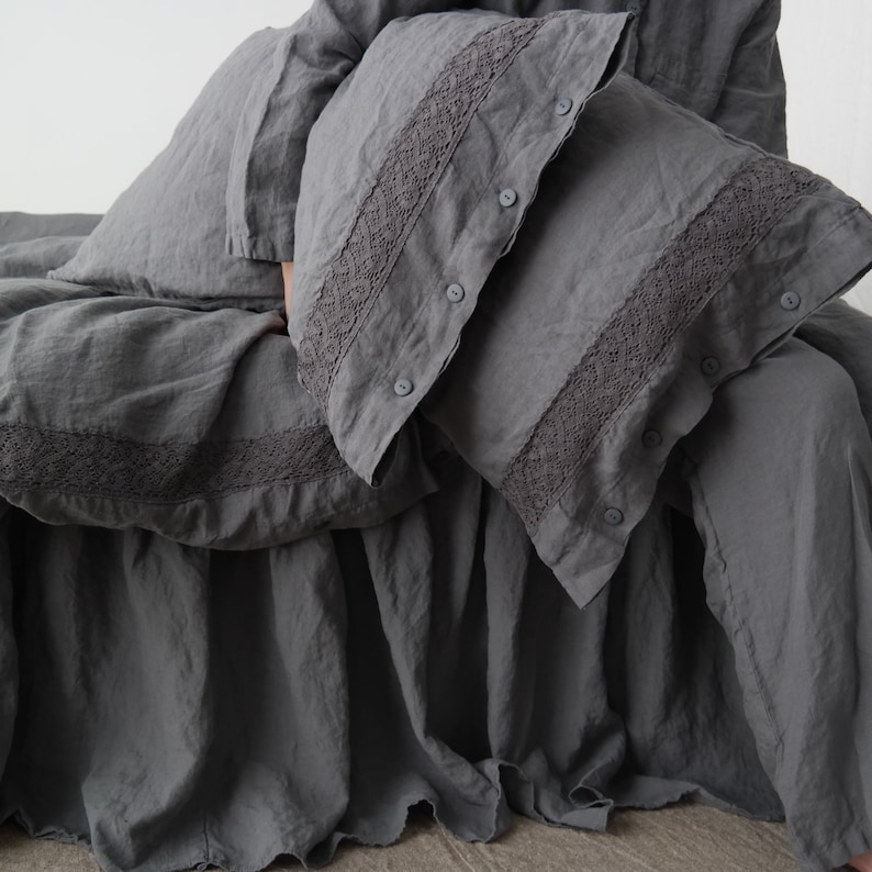 May include: A set of gray linen bedding, including a duvet cover, pillowcases, and a bed skirt. The pillowcases and duvet cover feature a decorative lace trim. The bedding is made of a soft, textured fabric and has a relaxed, casual look.