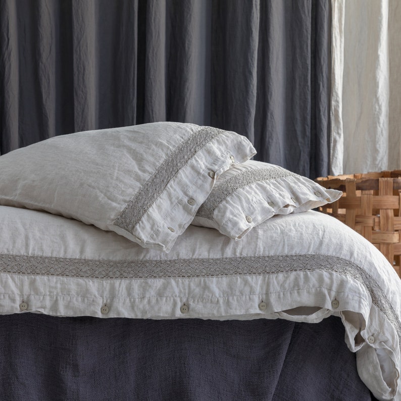 May include: A close-up of a bed with a cream-colored duvet cover and two matching pillowcases. The bedding has a gray embroidered trim and is buttoned. The bed is covered with a dark gray bed skirt. The background includes gray curtains and a woven basket.