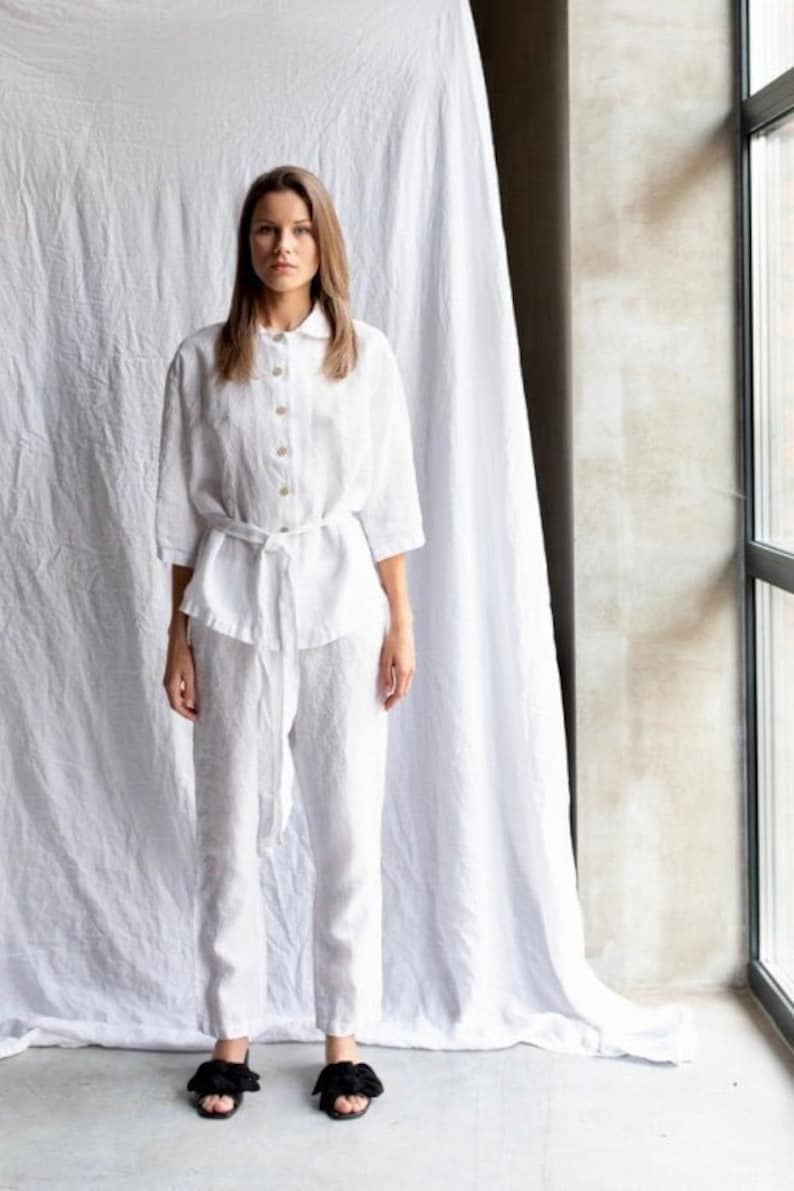 May include: A white linen shirt and pants set with a self-tie belt. The shirt has a collar and button closure. The pants have a relaxed fit.
