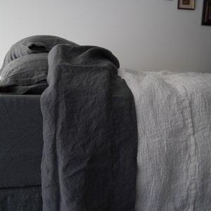 Puede incluir: Una cama con ropa de cama y almohadas de color gris carb&oacute;n oscuro superpuestas con una manta blanca texturizada. La tela gris oscuro tiene un aspecto liso y mate, mientras que la manta blanca muestra una textura tejida y arrugada.