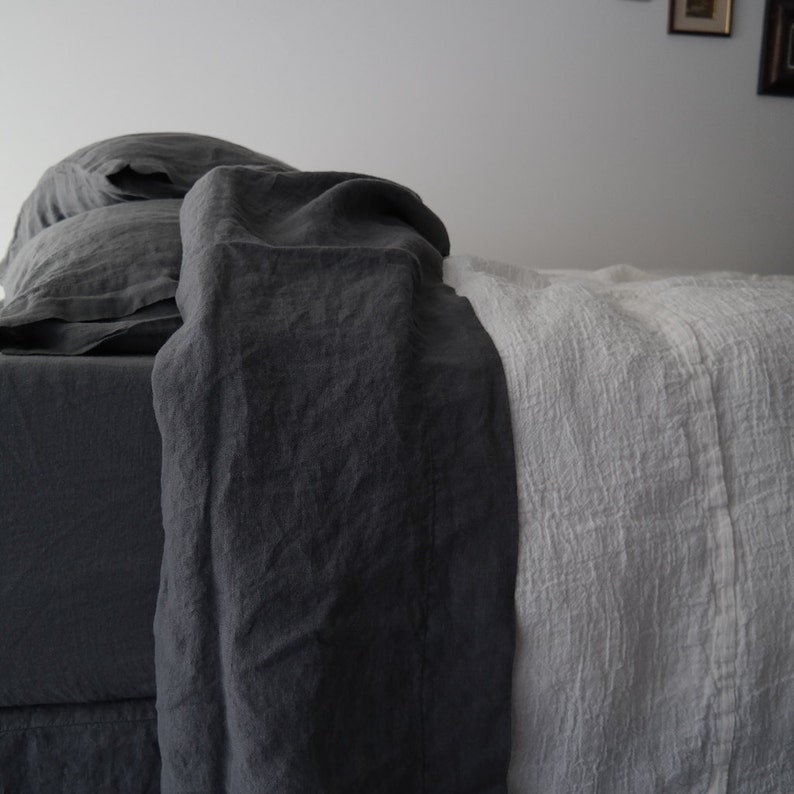 Puede incluir: Una cama con ropa de cama y almohadas de color gris carb&oacute;n oscuro superpuestas con una manta blanca texturizada. La tela gris oscuro tiene un aspecto liso y mate, mientras que la manta blanca muestra una textura tejida y arrugada.